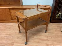 Full frontal view of teak bar cart on caster wheels with glass top insert and lower wooden shelf, showing general condition and wear.