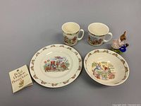 Set of two mugs, one bowl, one dinner plate, Dollie Bunnykins figurine and Royal Doulton Bunnykins booklet arranged on grey background.