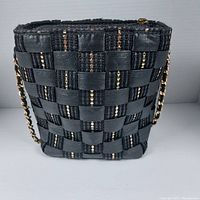 Front view of black and gold woven handbag with gold chain strap on white background.