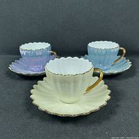 Three demitasse cups with matching saucers in cream, light blue, and pale lavender colors, each with gold trim and scalloped edges, displayed on black fabric background.
