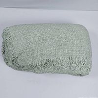 Folded light green woven blanket/throw with fringe on one edge showing texture and fabric detail.