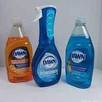 Front view of three dish soap bottles: Dawn Platinum Powerwash spray bottle with blue liquid, Dawn Ultra orange soap bottle, and Dawn Ultra blue soap bottle.