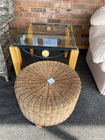 Photo showing square glass-top table with wooden legs and round wicker ottoman placed in front of brick wall and next to beige sofa.