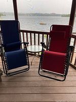 Two anti gravity lawn chairs, one in blue and one in red, placed on an outdoor deck by the water, with a small round metal glass-top table between them.
