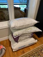 Stack of 4 bedding items including duvets and pillows wrapped in plastic, placed on wood floor near window and cabinet.