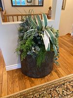 Front and angled view of faux plant with various green and white leaves in a large round brown container sitting on hardwood floor next to a small railing.