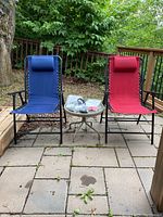 Photo showing two folding lawn chairs - one red and one blue - placed on outdoor patio, with small round metal table between holding first aid items.