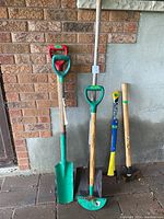 Photo showing five gardening tools including shovel, two shovel edgers, blue/yellow axe, wooden axe, all placed upright against brick and grey wall.