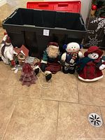 Black plastic storage tote behind holiday decorations including stuffed snowmen, a Santa figure, ceramic house, and small tree figurine on tile floor.