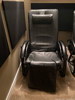 Front view of black leather-like massage chair with extended footrest and cushioned headrest in a carpeted room with soundproofing panels.
