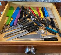 Top-down view of a drawer filled with various kitchen knives, peelers, and measuring spoons arranged with different colored handles and protective sheaths on knives.