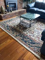 View showing full carpet with floral and leaf motifs bordered by dark trim, placed on hardwood floor under glass coffee table.