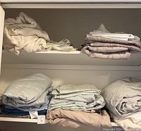 Shelves containing folded and rolled assorted bed sheets including neutral tones and light lavender patterned sheets.