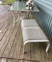Long shot showing rectangular glass-top table with white metal frame, two metal mesh ottoman benches, all placed on wooden deck.