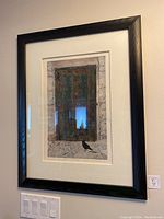 Framed picture of a rustic window with green shutters and a black bird on ledge, hung on a wall above light switches.