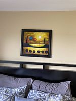 Framed print hanging on beige wall above black bed headboard, showing square art with stylized trees, sun and reflection.