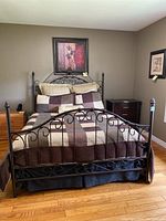 Front view of queen size metal bed frame with brown and beige patchwork quilt and several pillows arranged on bed in a room with hardwood flooring and wall decor.