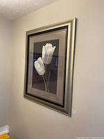Framed print showing two white tulips with a silver and black frame hung on a wall. The image shows the overall view of the artwork in a setting.