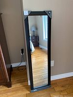 Full view of the rectangular wall mirror with black frame leaning against a beige wall, showing the reflective surface and wooden floor.