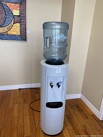 Front view of white water cooler with transparent blue water jug on top, showing two dispensing taps.