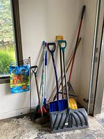 Various winter shovels, a rake, and brooms arranged against garage wall with a decorative welcome flag on stand to the side.
