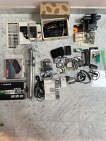 Full overview of all items in lot, including cameras, charger, tripod, cassette recorder, battery, cords, flash cubes, and boxes.