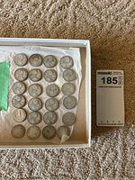 Photo showing 20 coins including 19 Canadian quarters and one American quarter arranged in rows inside a box with tissue paper.
