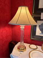 Full view of the entire lamp showing the beige fabric shade, crystal column, and polished brass base on a white cloth.