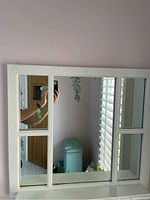 White window pane style wall mirror hanging on a light purple painted wall. Four narrow rectangular vertical mirrors on the sides and a large rectangular mirror below serve as a display shelf.