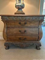 Front view of wooden side table with three drawers and metal handles. Decorative inlay on top surface and carved detailing under the edges.