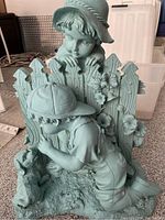 Full view of teal resin statue of a little boy kneeling by a dog in front of a wooden picket fence with a little girl leaning over the fence behind him.