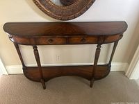 Front view of half-moon shaped wooden hall table with two center drawers and lower shelf under a wall mirror.