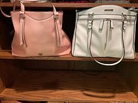Two leather handbags on a wooden shelf: beige Simply Noelle bag on left and white handbag with black trim on right.