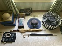 Photo of multiple ladies hats displayed on floor. Includes beige hat in box, blue-grey hat, black and white patterned hats in striped round boxes, a small metallic purse, umbrellas, jewelry box, belt and folded fabric garment bags.