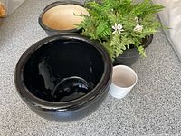 Top view of three large black ceramic planters (two with beige interiors visible) and one small white planter beside an artificial green fern plant with white flowers inside one large planter.