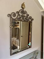 Full view of the rectangular mirror mounted on a wall, showing the textured metal frame with intricate scroll patterns and leaf finial.