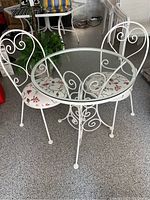 White metal patio bistro set with round glass top table and two matching white metal chairs with floral upholstered seats, showing front angle.