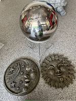 Three items shown together: reflective gazing ball on stand, crescent moon and stars wall sculpture, sun wall sculpture.