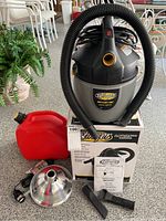 Full lot showing wet/dry vacuum, flexible hose coiled over vacuum, plastic red gas can, clamp lamp with reflector, two nozzle attachments, and owner's manual.