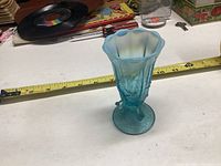Blue opalescent glass bud vase on a white surface with measuring tape showing about 6 inches.