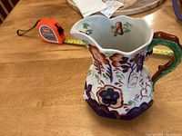 Front angle of the ceramic jug showing the snake handle and floral decoration on a wooden table with measuring tape illustrating size.