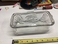 Clear rectangular glass refrigerator dish with vegetable embossed lid shown on white surface with measuring tape.