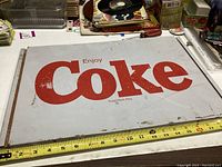 Metal rectangular sign with 'Enjoy Coke' written in red on a white background, showing wear and scratches, measuring approximately 18 inches wide.