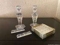 Pair of cross pattern crystal candlesticks with two crystal cutlery rests and an unopened box likely containing more rests, displayed on reflective surface against beige background.