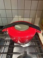 Side top-angle view of red cast enamel pot with glass lid and black handle placed on stovetop.
