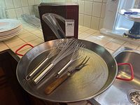 Photo showing metal paella pan holding three wire whisks and one fork with wooden handle, as well as the tiled kitchen background and part of white plates.