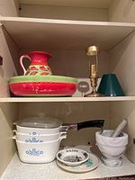Shelf with red and green ceramic bowl and jug, brass candleholder with green shade, white Corning Ware dishes, small plates, and white marble mortar and pestle.