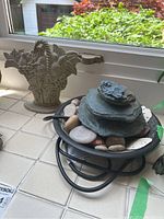 View of both cast metal door stop and electric tabletop water feature on a windowsill. Door stop shows basket of fruit motif, water feature has stacked stone design with electrical cord.