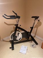 Full side view of the black stationary exercise bike showing frame, flywheel casing, seat, handlebars, and user manual on the floor.