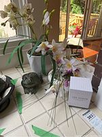 View of two faux orchids in white pots on a tiled windowsill, metal frog figurine in front, and boxed candle labeled The North beside them.